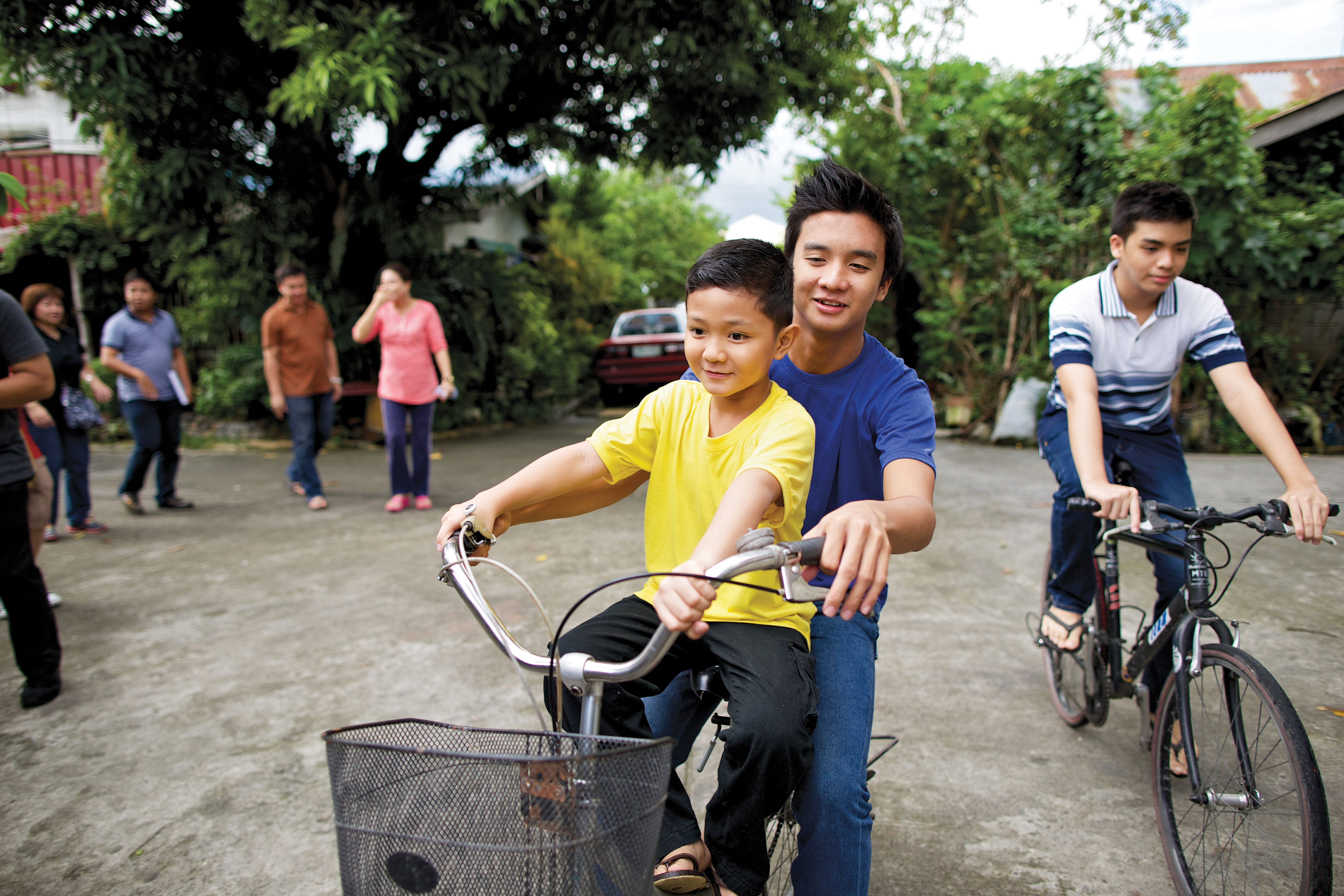 A young boy in a yellow T-shirt rides on a bike seat in front of his older brother in a blue T-shirt, both holding the handlebars.