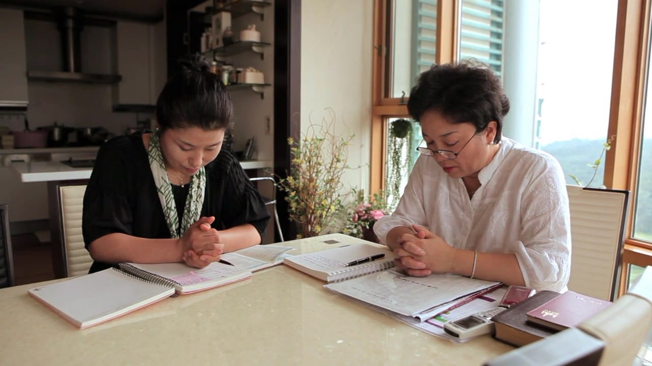 Two women pray together at a table with binders