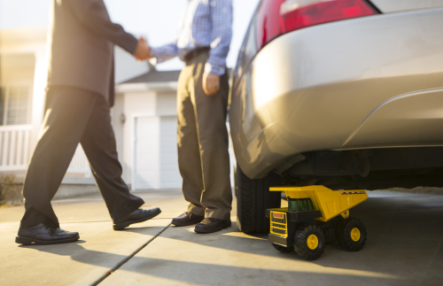 Two men are shaking hands in a driveway by a car.  There is a yellow toy truck behind the back wheel of the car.
