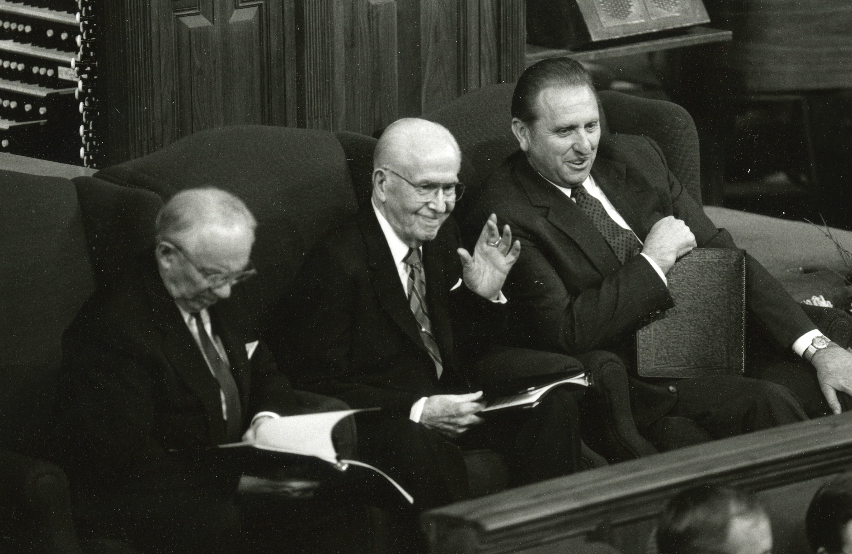 President Benson sitting in a large armchair between his counselors, smiling and waving to the audience in the Tabernacle.
