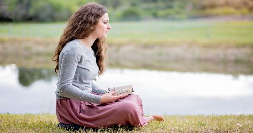 Young adult woman ponders scriptures near lake.