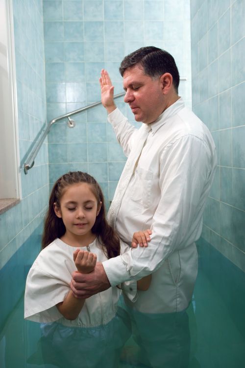 A girl with long brown hair, wearing a white jumpsuit, being baptized in a baptismal font by a man in a white shirt and tie.