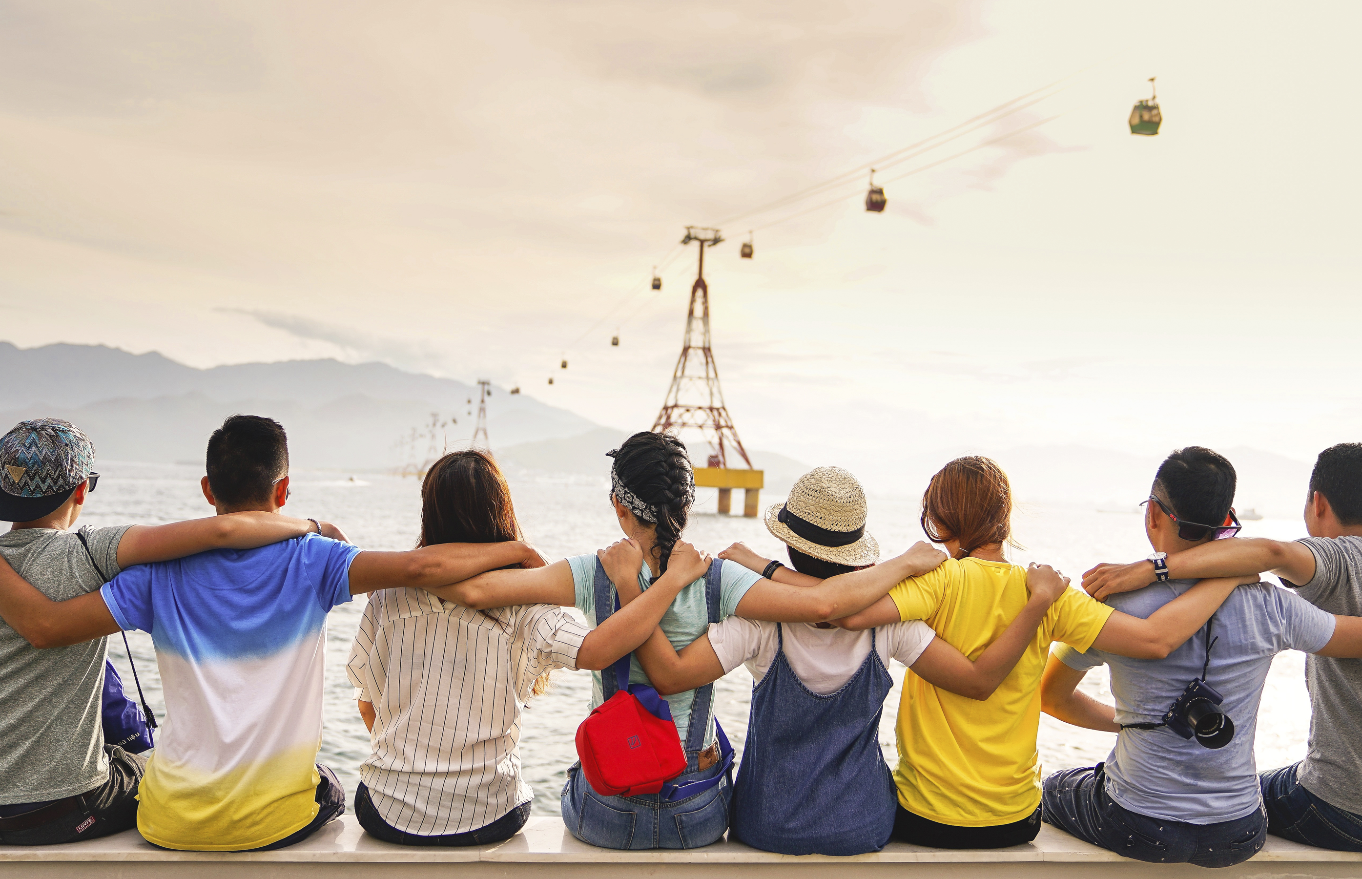 Teenagers are sitting on a wall facing the ocean. Their backs are to the camera and they are their arms over one each others shoulders.