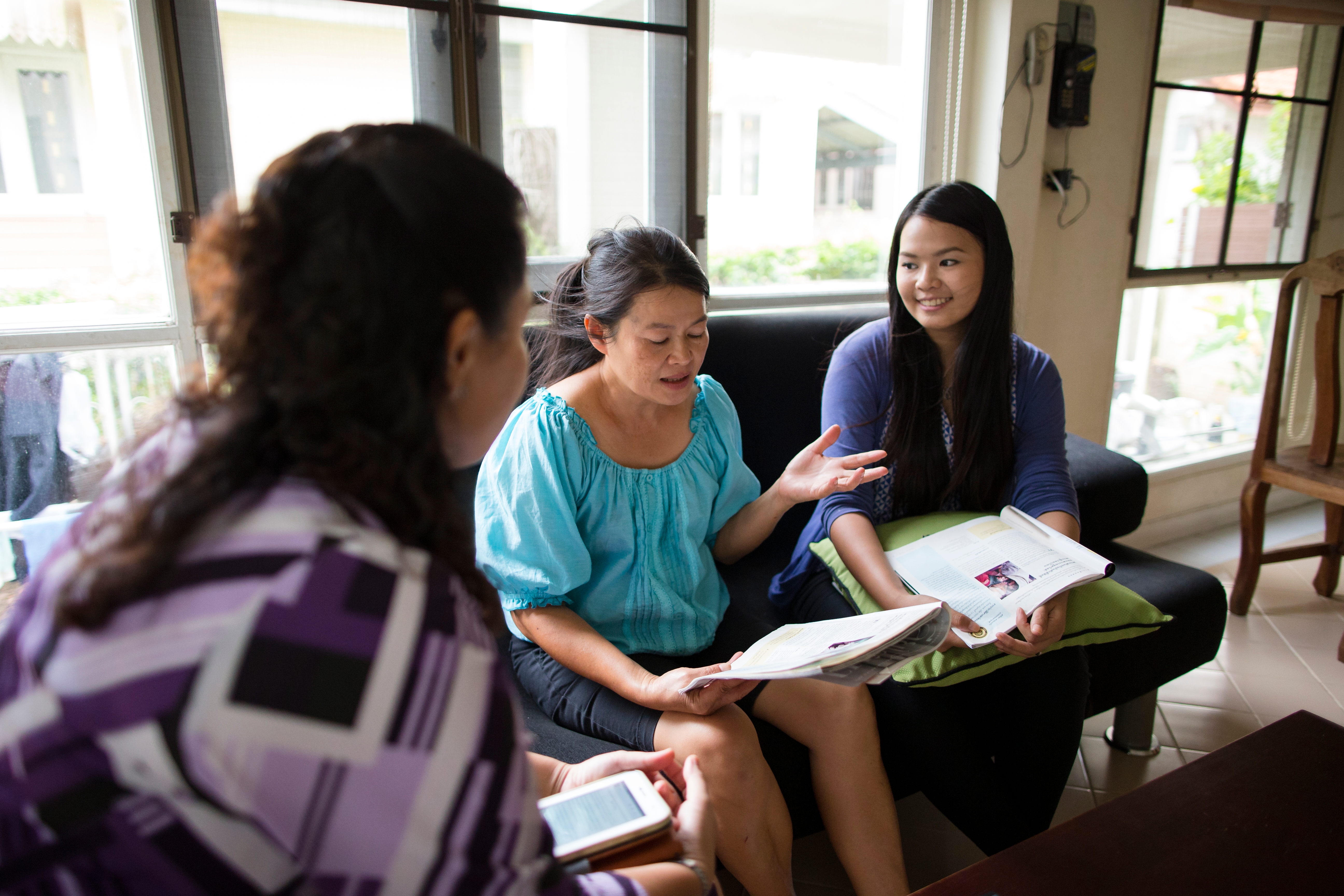 A woman in a blue shirt sitting on a couch and reading from a magazine with two women on either side of her, studying together.