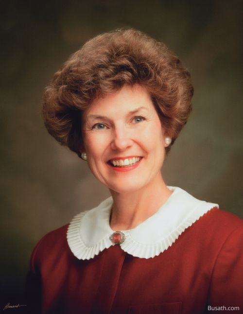 A photograph of Barbara Woodhead Winder against a gray background, wearing a red dress with a white collar.