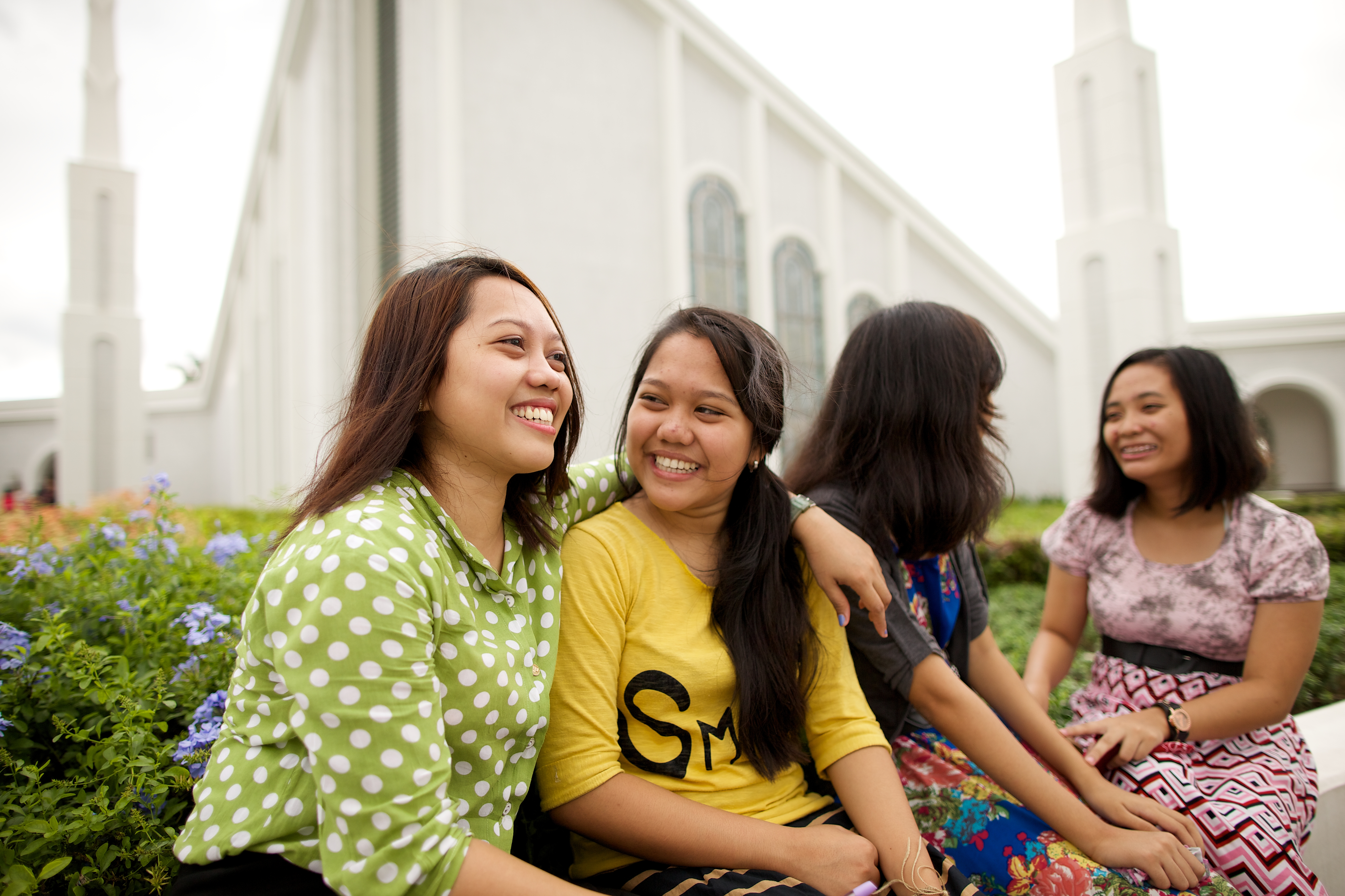 A young woman putting her arm around another young woman’s shoulders, with two other women sitting nearby outside the Manila Philippines Temple.