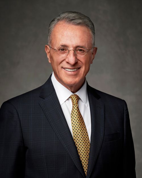 A portrait of Elder Ulisses Soares wearing a black suit and a red and blue striped tie.