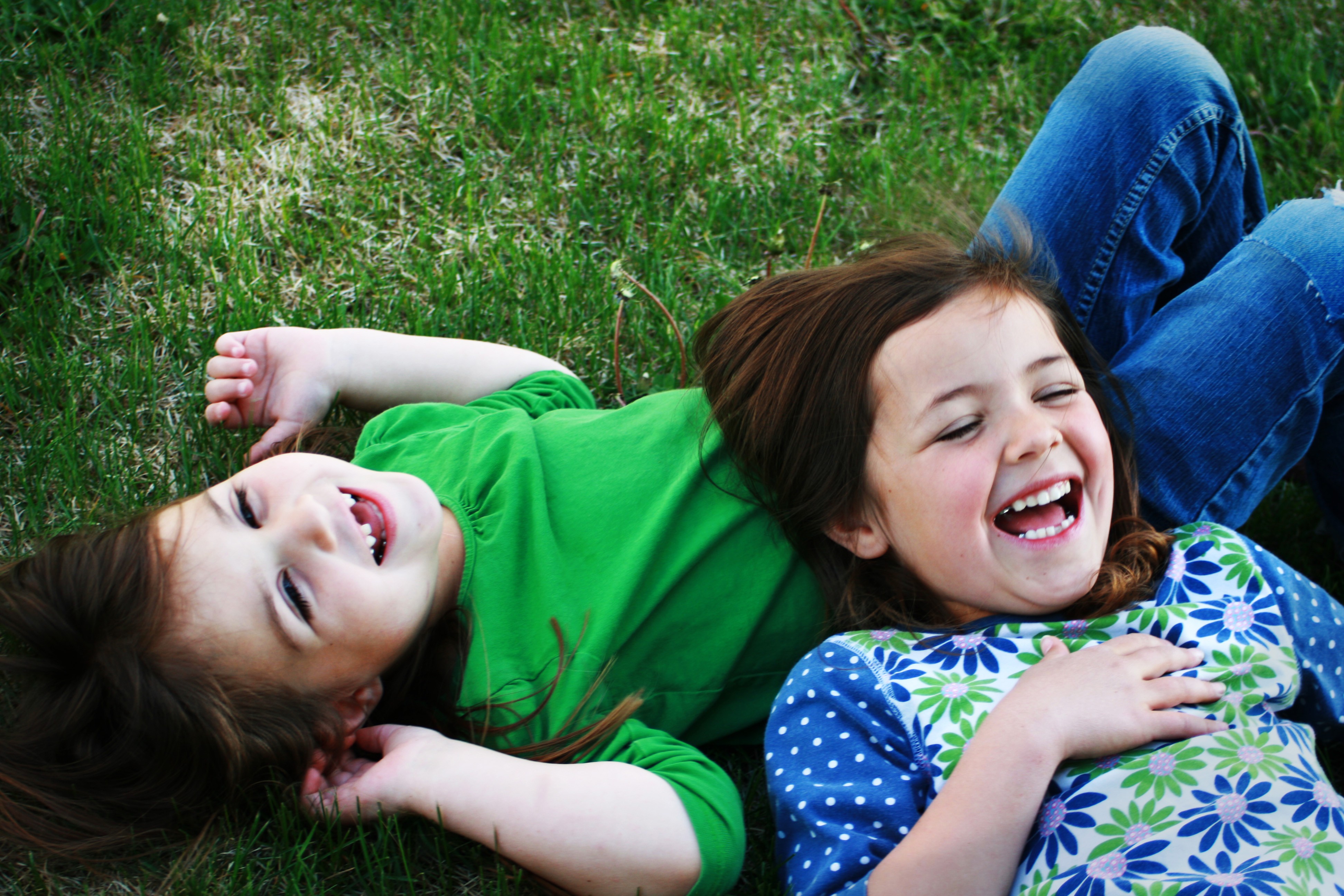 A girl lying in the grass with another girl lying on her stomach as both laugh.