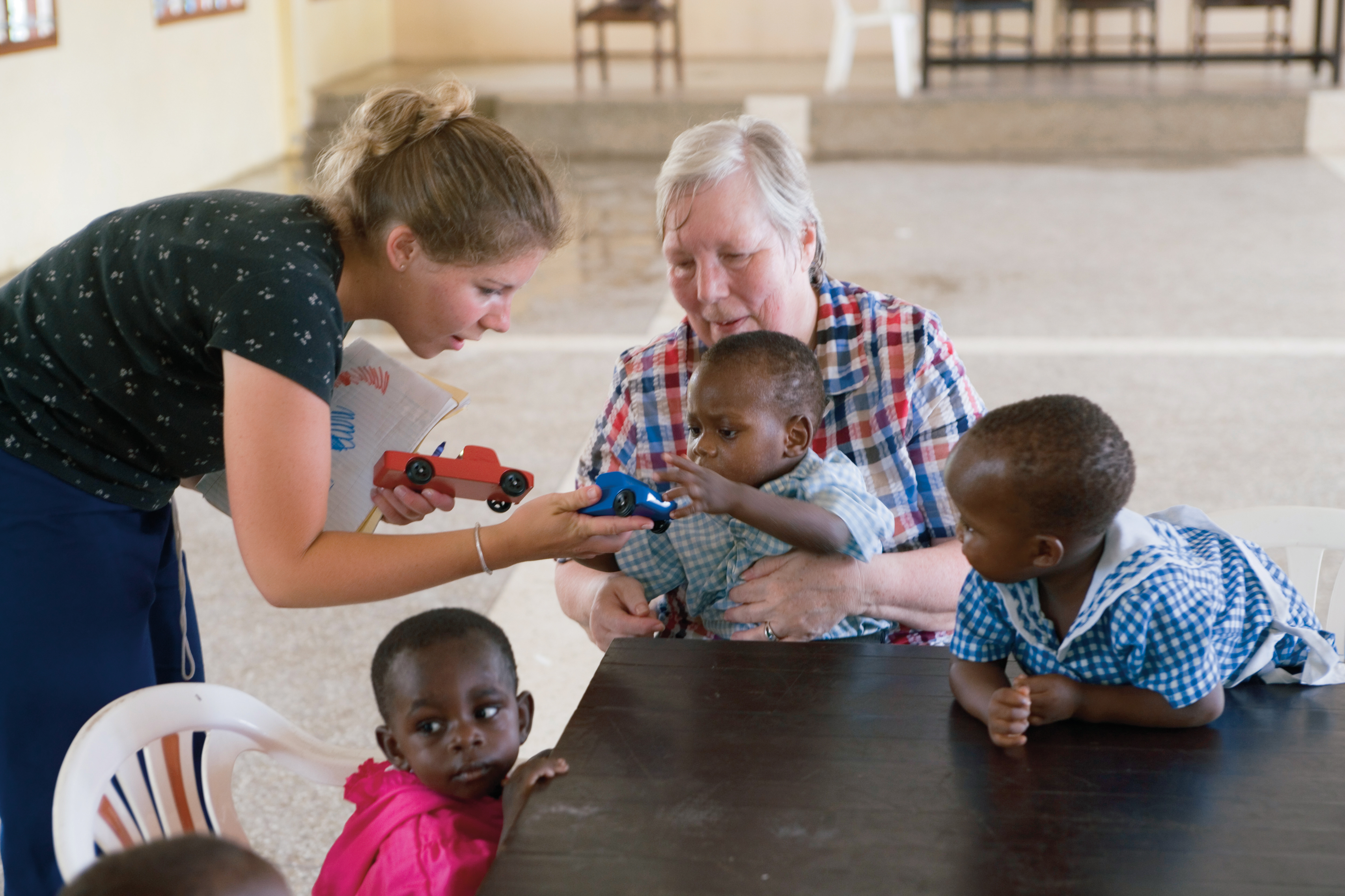 A woman in an orphanage holds an African child while another woman hands the child a wooden car, with two other children nearby.