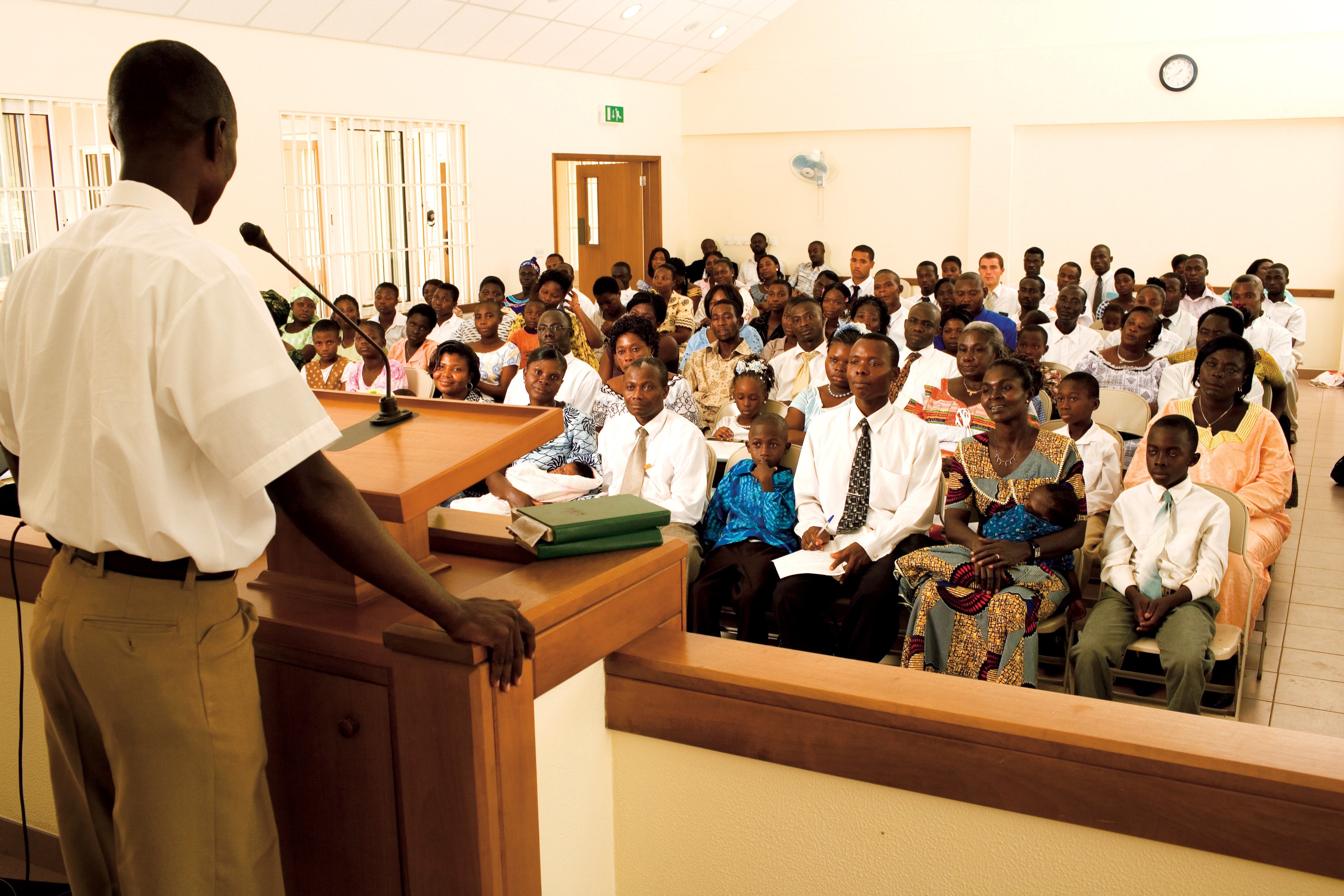 A man in a white shirt stands in front of a large congregation in Ghana and delivers a talk.
