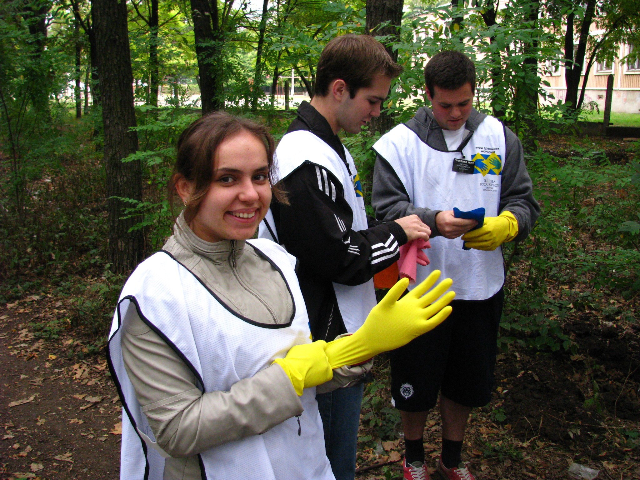 Two young men and a young woman in Mormon Helping Hands vests standing outside and putting on rubber gloves.