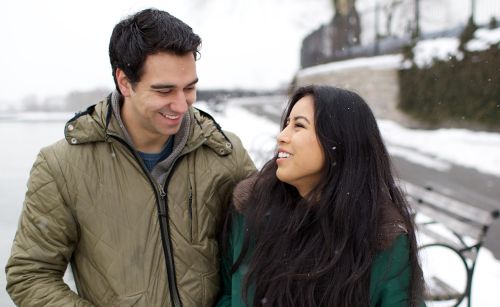 A young man and woman are walking and talking together on a sidewalk surrounded by snow in New York City.  They are also stopped looking our over a lake or river.