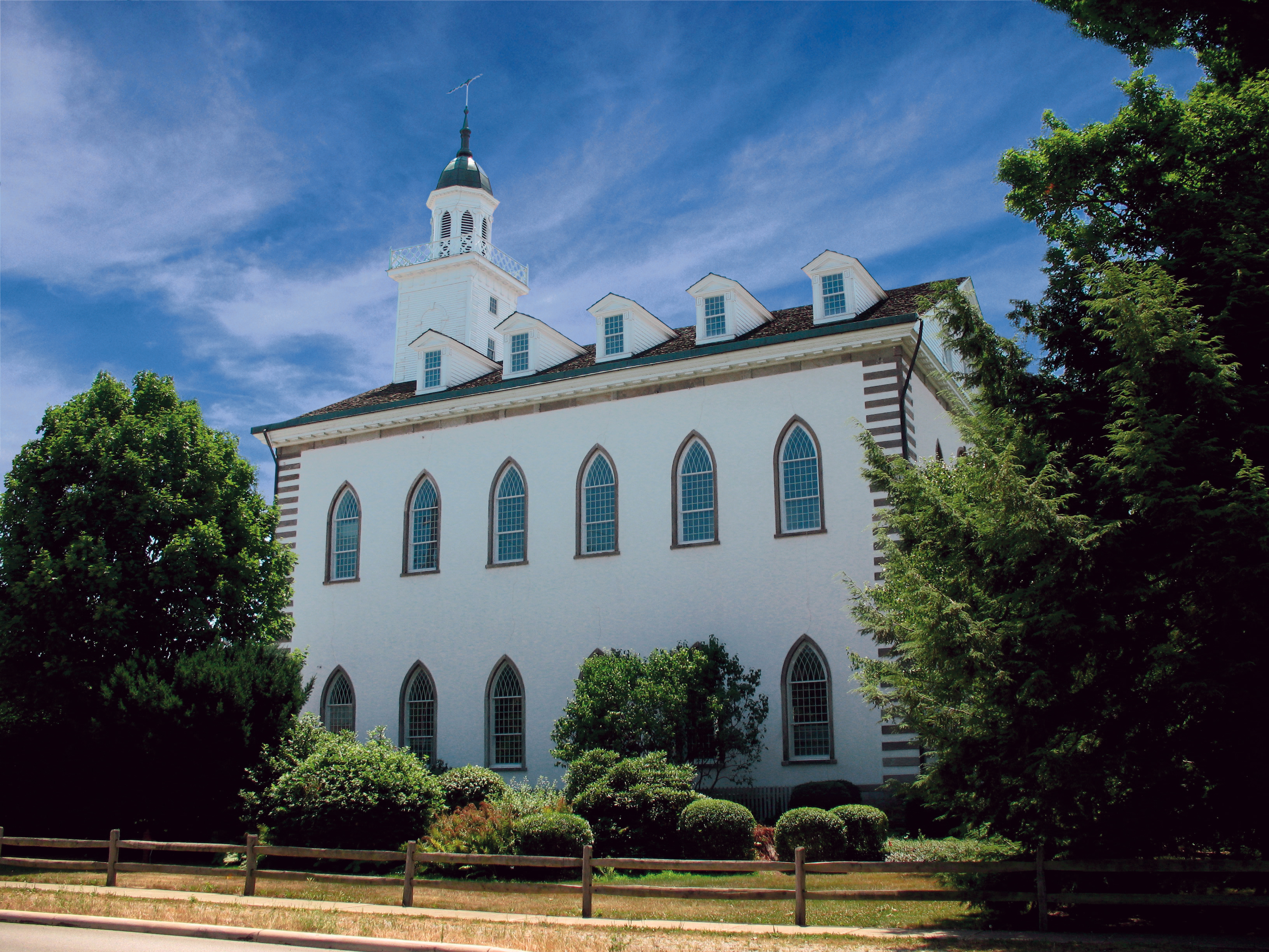 The Kirtland Temple seen from the side between two large green trees, with a small wooden fence running around the grounds.