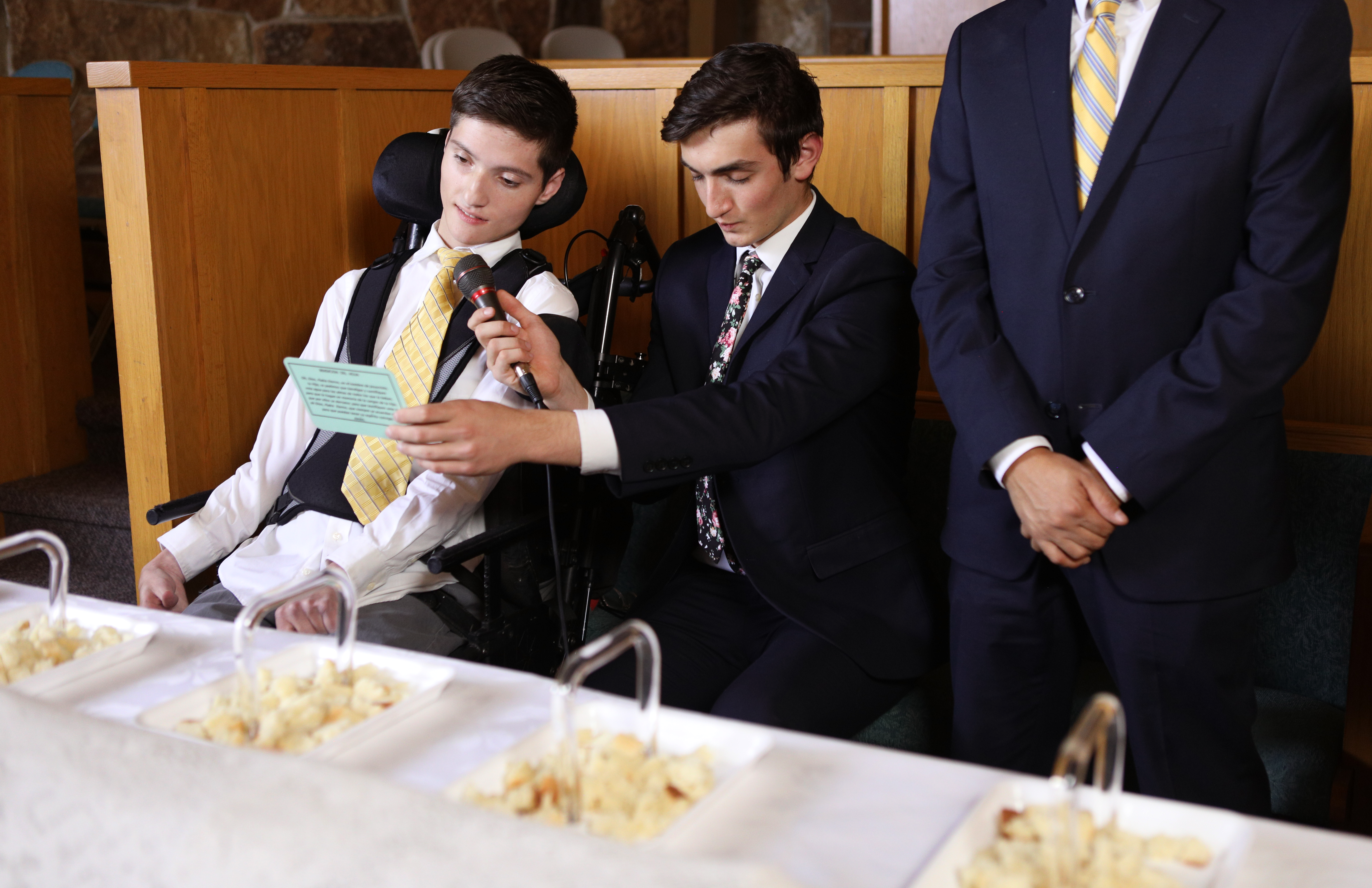 A young man who is in a wheelchair has a friend next to him holding the sacrament prayer and microphone so he may bless the sacrament.