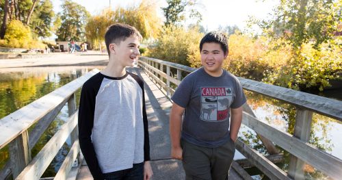 橋を渡る二人の青少年。秋の季節。男の子たちが会話をしている様子。湖の上にかかる橋。遠くに見える草原や遊具。