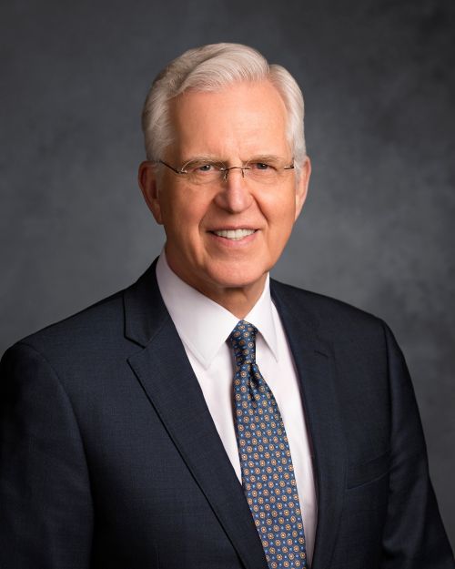 A portrait of Elder Neil L. Andersen, who is wearing a black suit and a solid red tie, in front of a gray background.