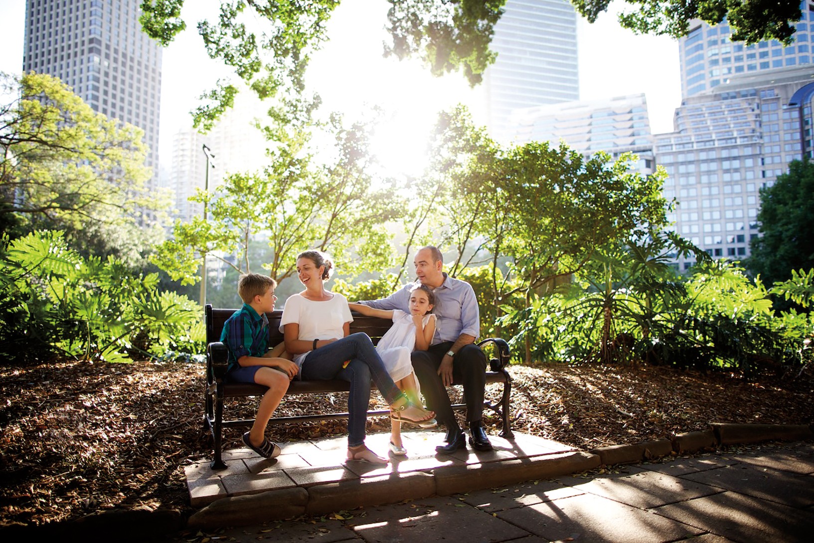 A mother and father sit on a bench in a park with their two children.
