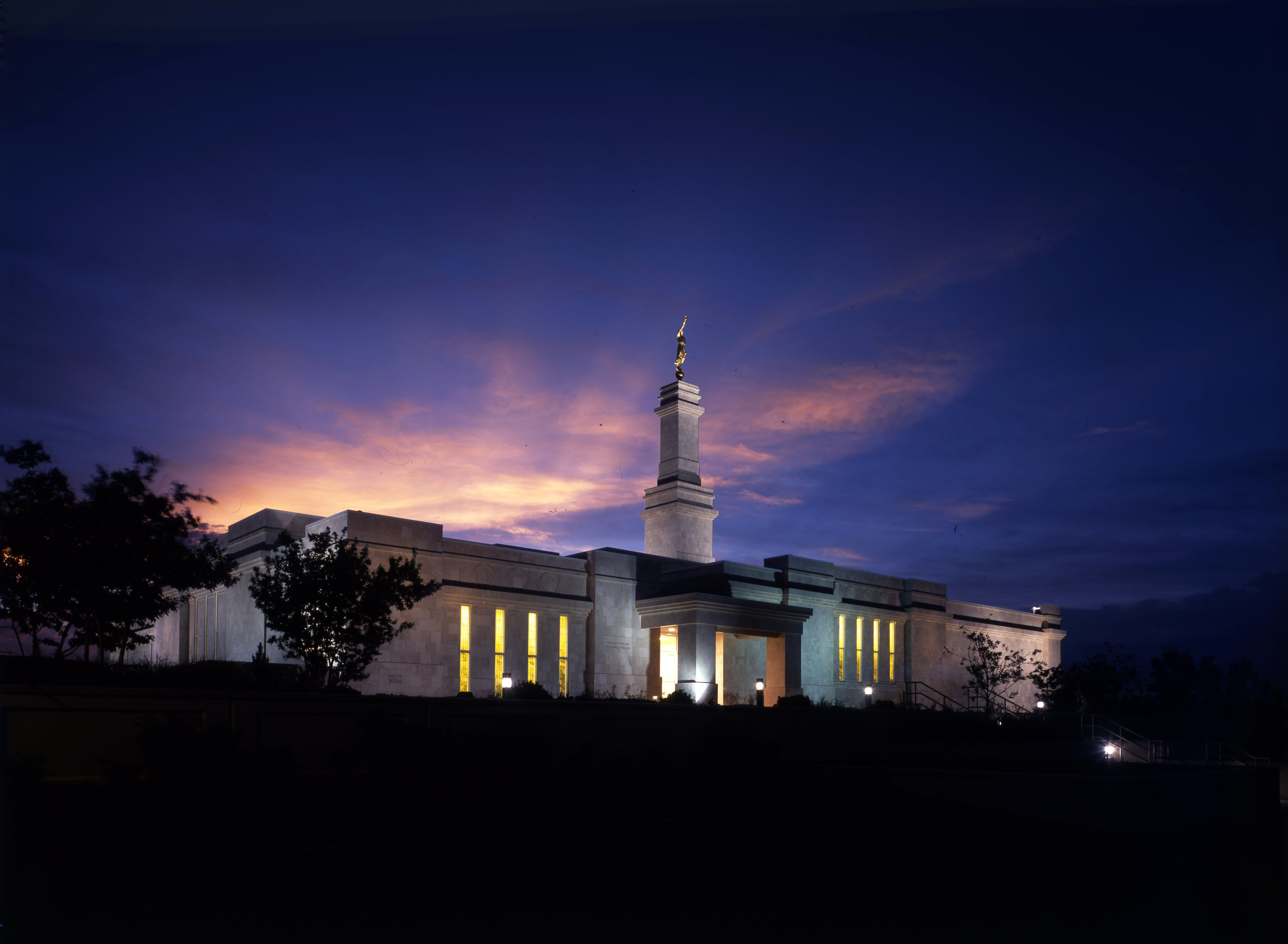 The Monticello Utah Temple is lit up with exterior lights in the evening, with scenery of silhouetted trees against a sunset sky.