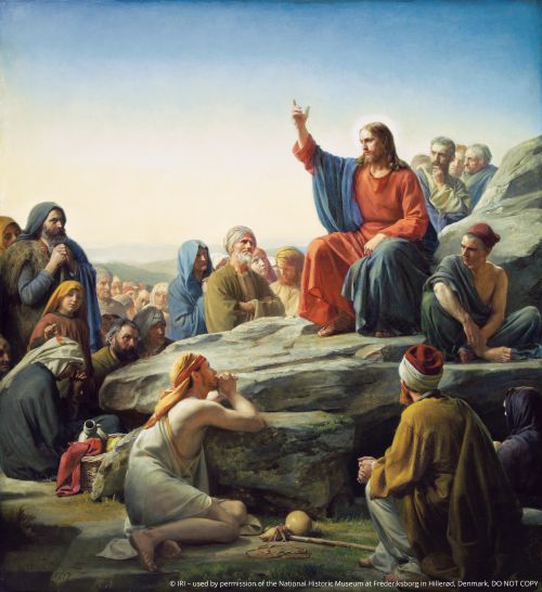 Christ in red and blue robes, sitting on a large rock and teaching, while a large group of people sit around Him listening to His words.