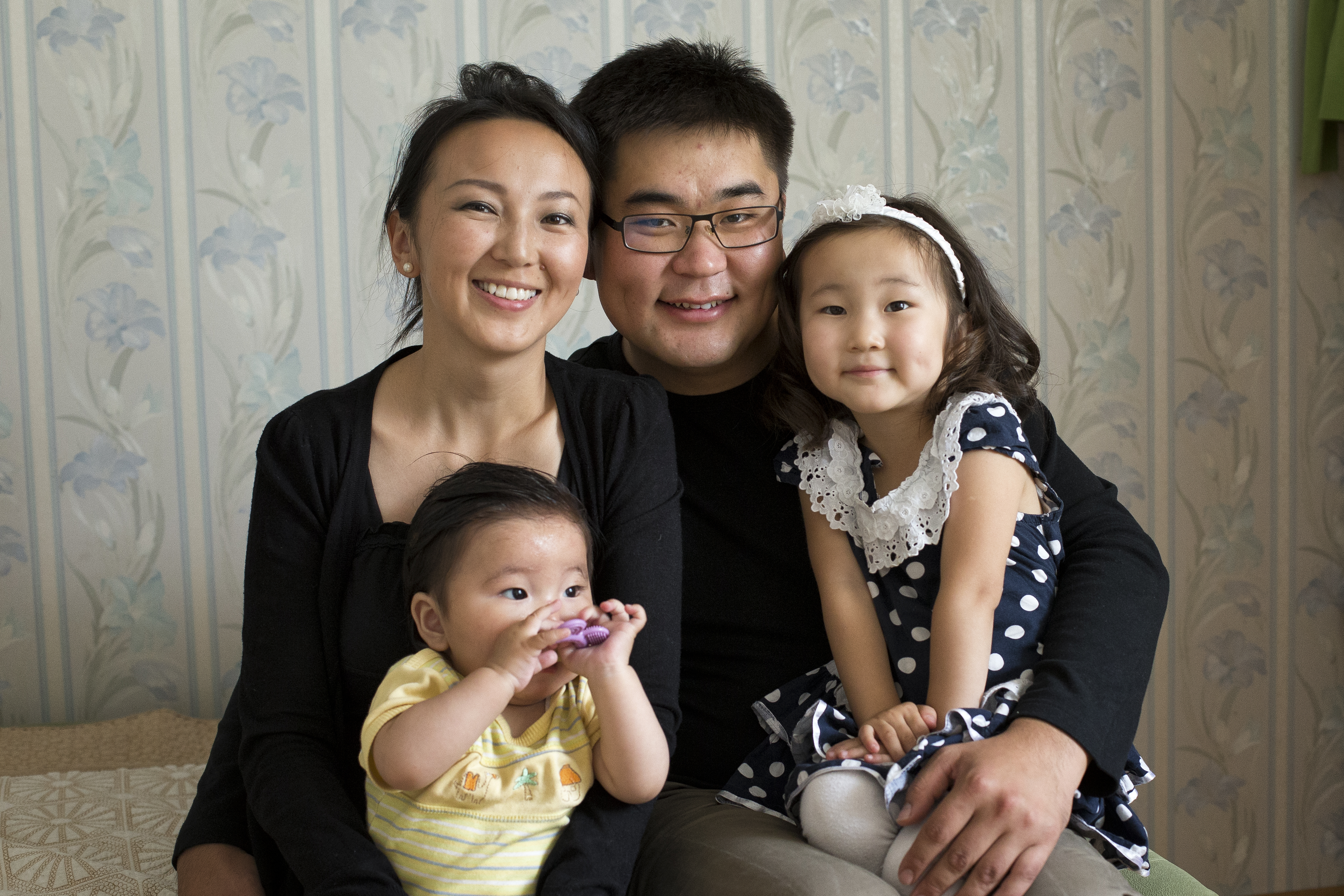 A husband and wife sit next to one another and smile with their two young children on their laps.