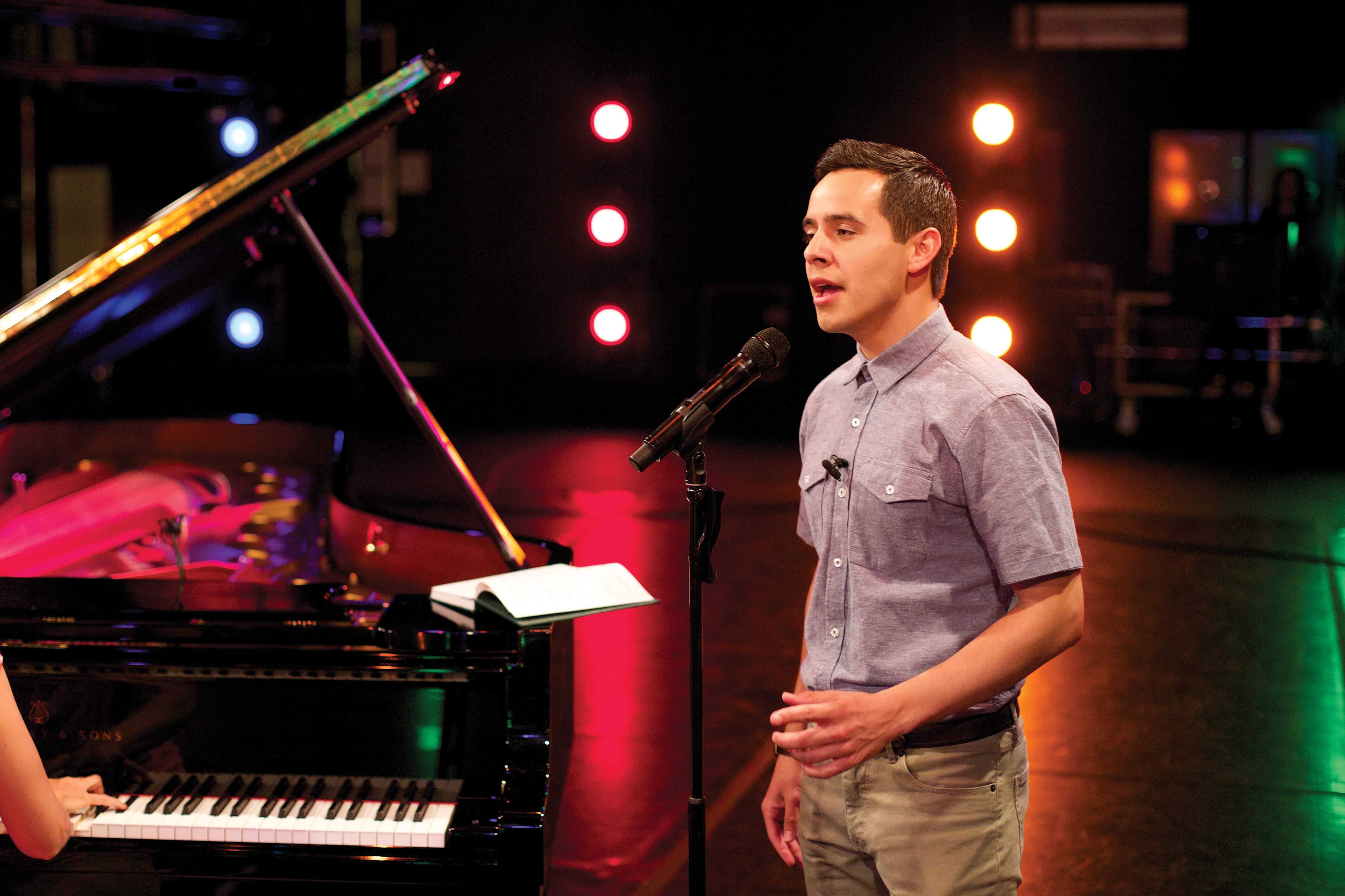 David Archuleta stands on a lighted stage and sings next to a piano.