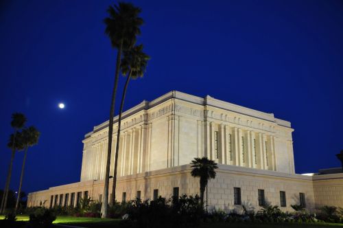 The Mesa Arizona Temple and palm trees lit up at night, with a dark blue sky and moon in the distance.