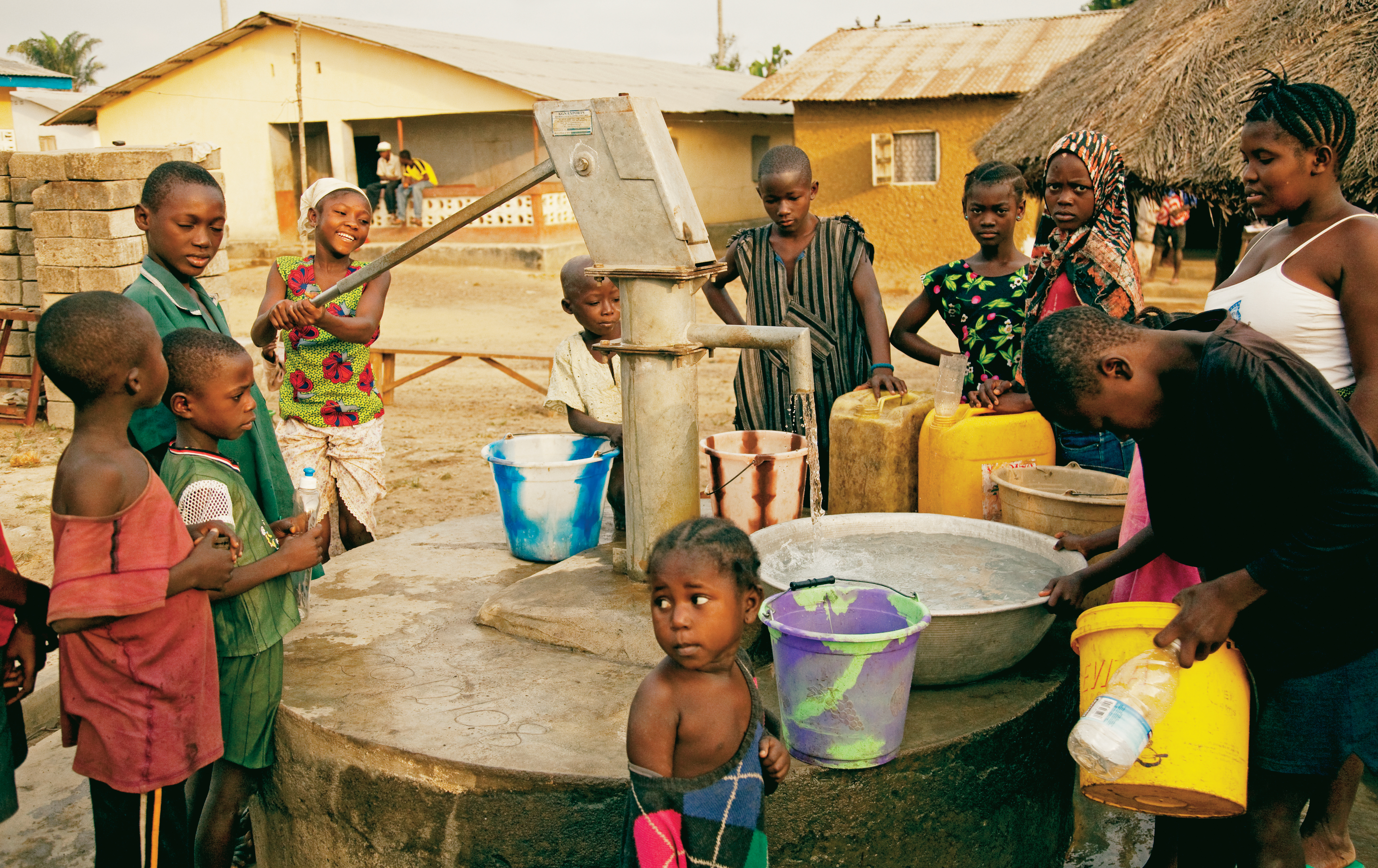 African children and teenagers stand around the hand-operated water pump while a young woman lifts the pump lever, filling a bucket with water.