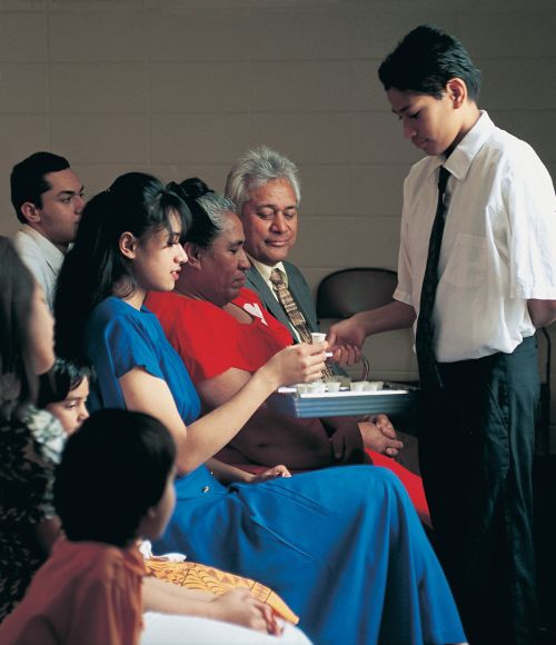 A deacon in a white shirt and tie is holding out the water sacrament tray to a young woman in a blue dress, who is holding a sacrament cup.