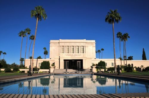 The Mesa Arizona Temple reflected in a pool on a sunny day, with surrounding palm trees and a blue sky.