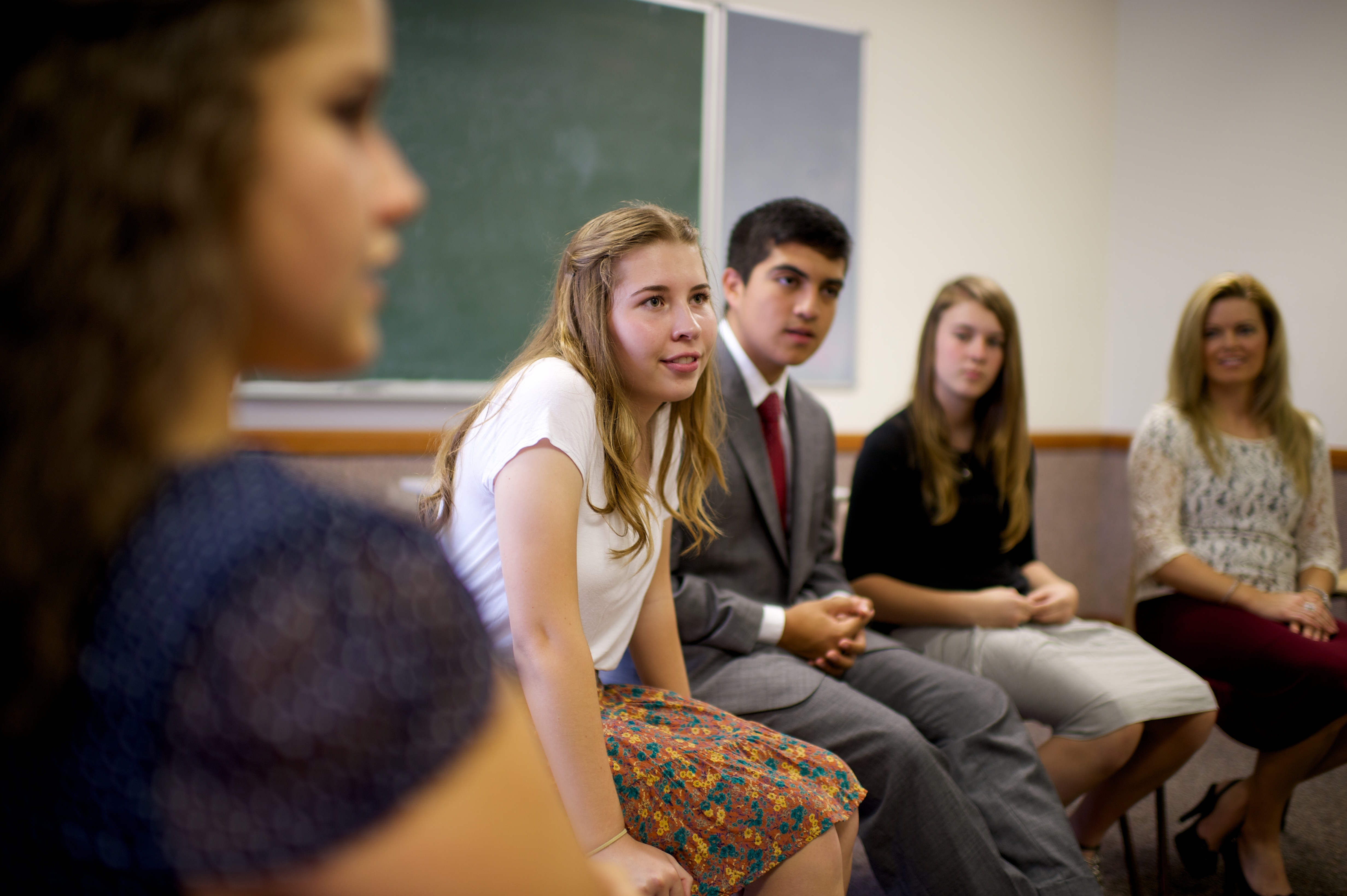 A young woman with long blond hair, wearing a skirt, leans forward in her chair and looks to the front of the classroom with other young men and women.