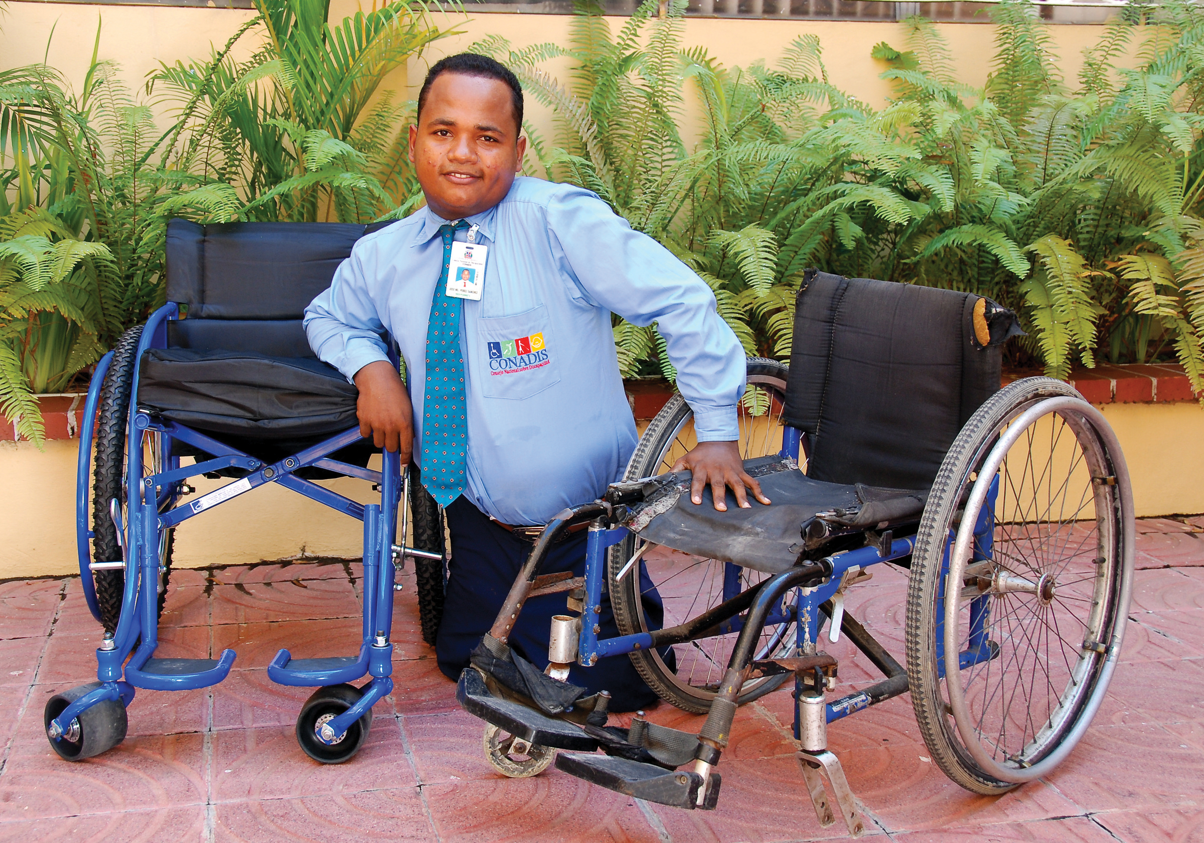 A man from the Dominican Republic balancing himself between a rusting, torn, dirty wheelchair and a new, shiny black and blue wheelchair.