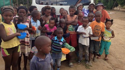 Jacques du Plessis stands with  number of children in Africa who all seem to be holding bowls. There is another man standing with them and there is a truck behind all of them.