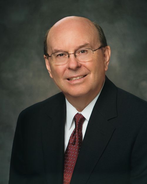 A portrait of Elder Quentin L. Cook, who is wearing a black suit and a maroon and white tie, sitting in front of a gray background.