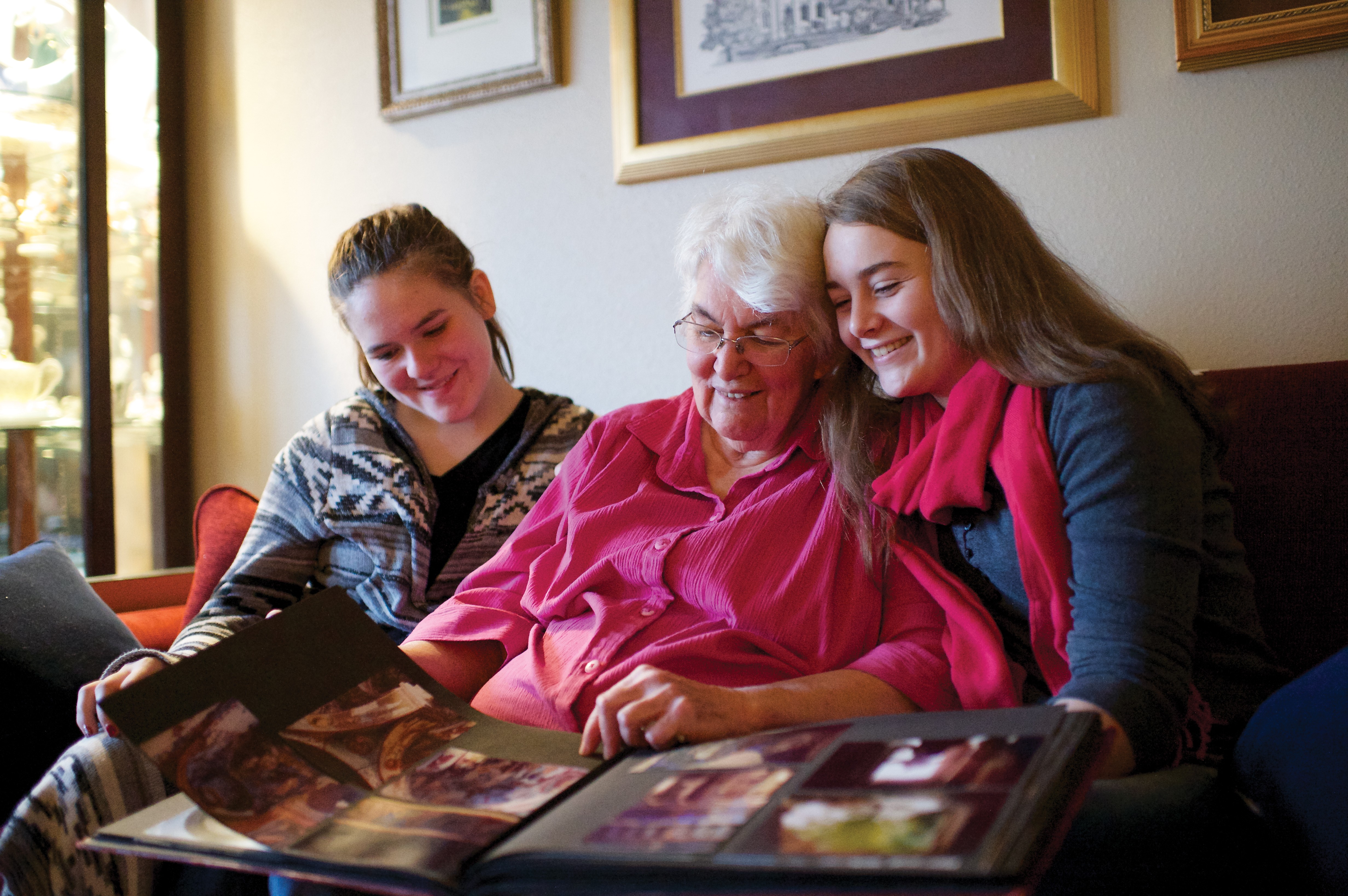 An elderly woman sits on a couch with her two granddaughters, looking at an album of family photographs.