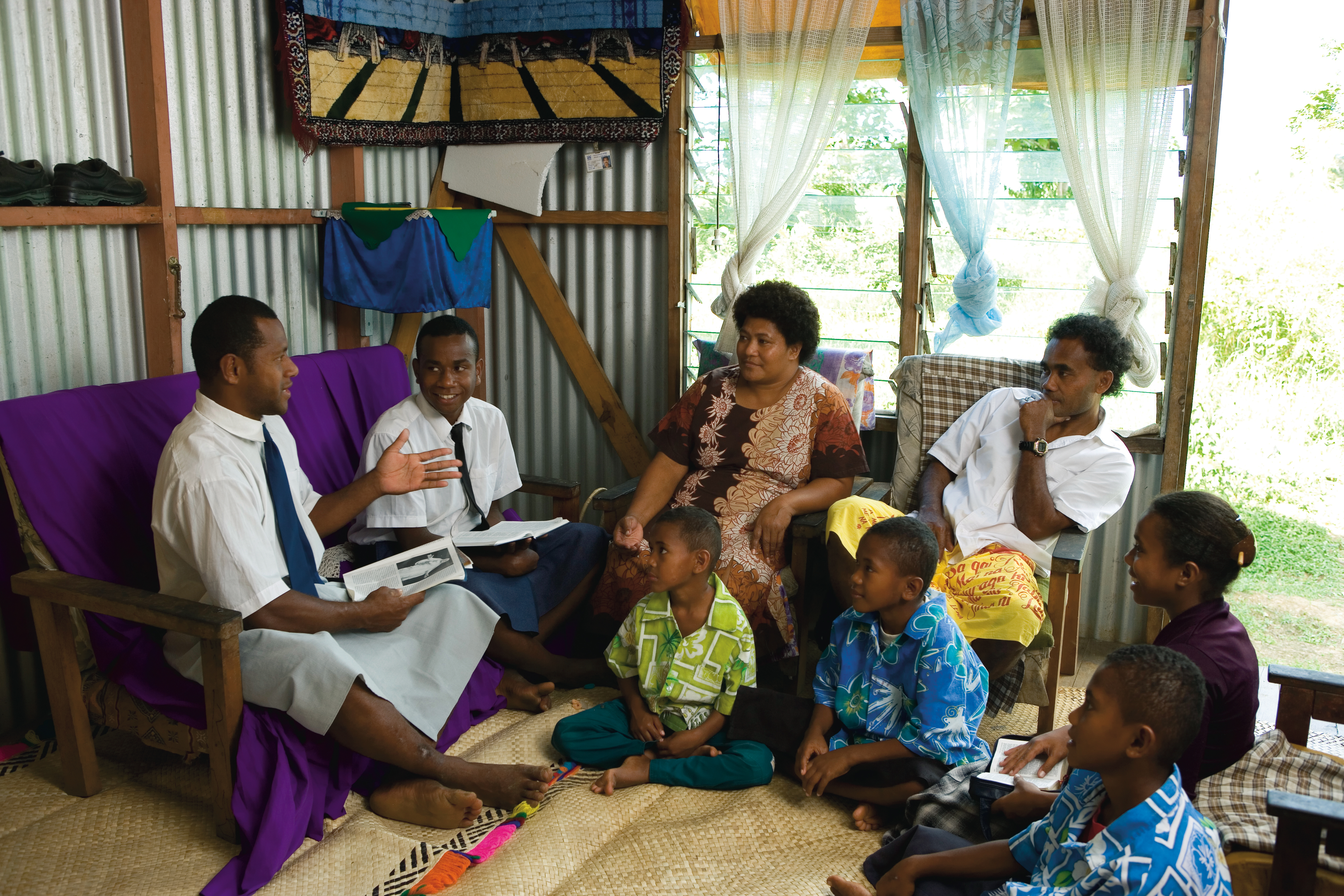 Two elder missionaries sit on a blue couch and teach a father, mother, and four children, who are sitting on chairs and the floor in their home in Tonga.