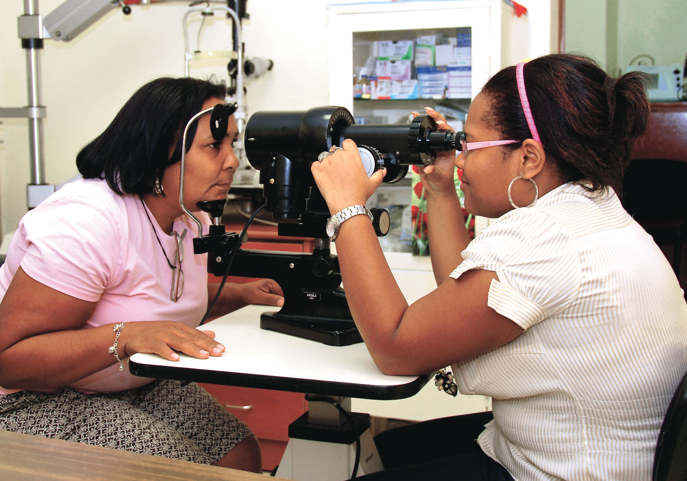 A Dominican woman in a light pink shirt sitting and resting her chin on an eye examination device while a doctor checks her eyesight.