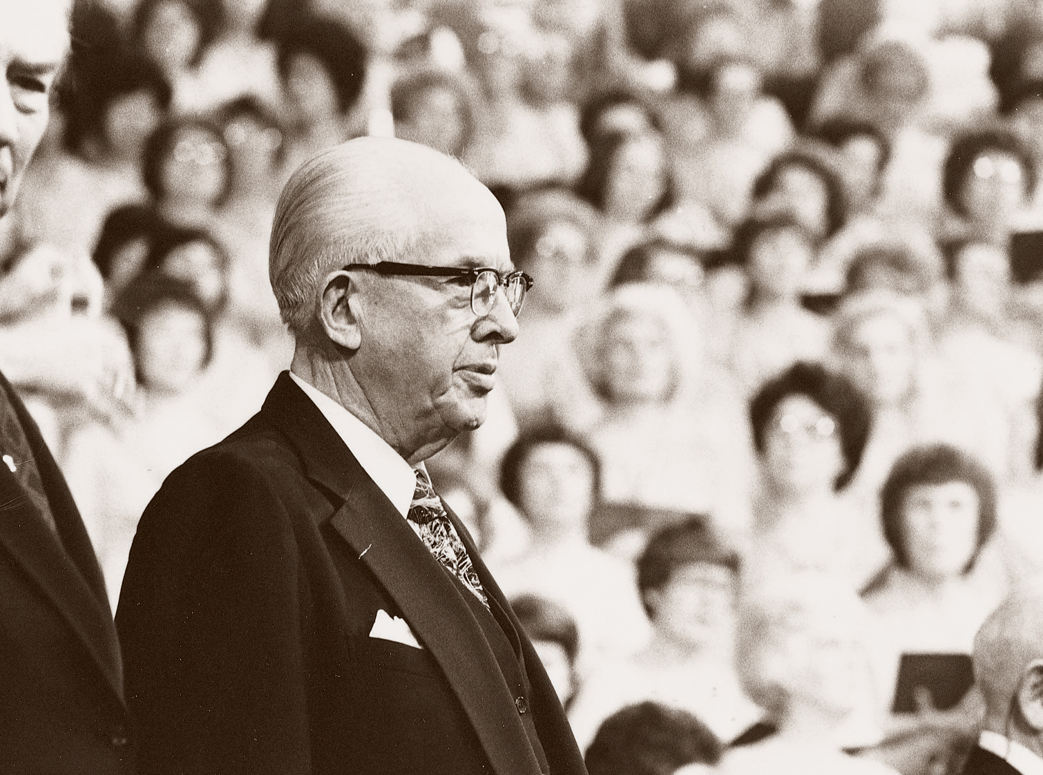 President Benson in a suit, a white shirt, and glasses, standing and speaking to the audience at general conference.