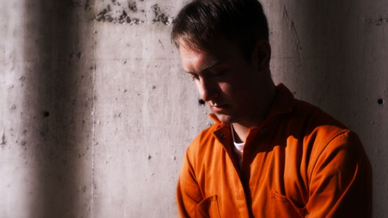 A man in an orange jumpsuit sits against a concrete wall.