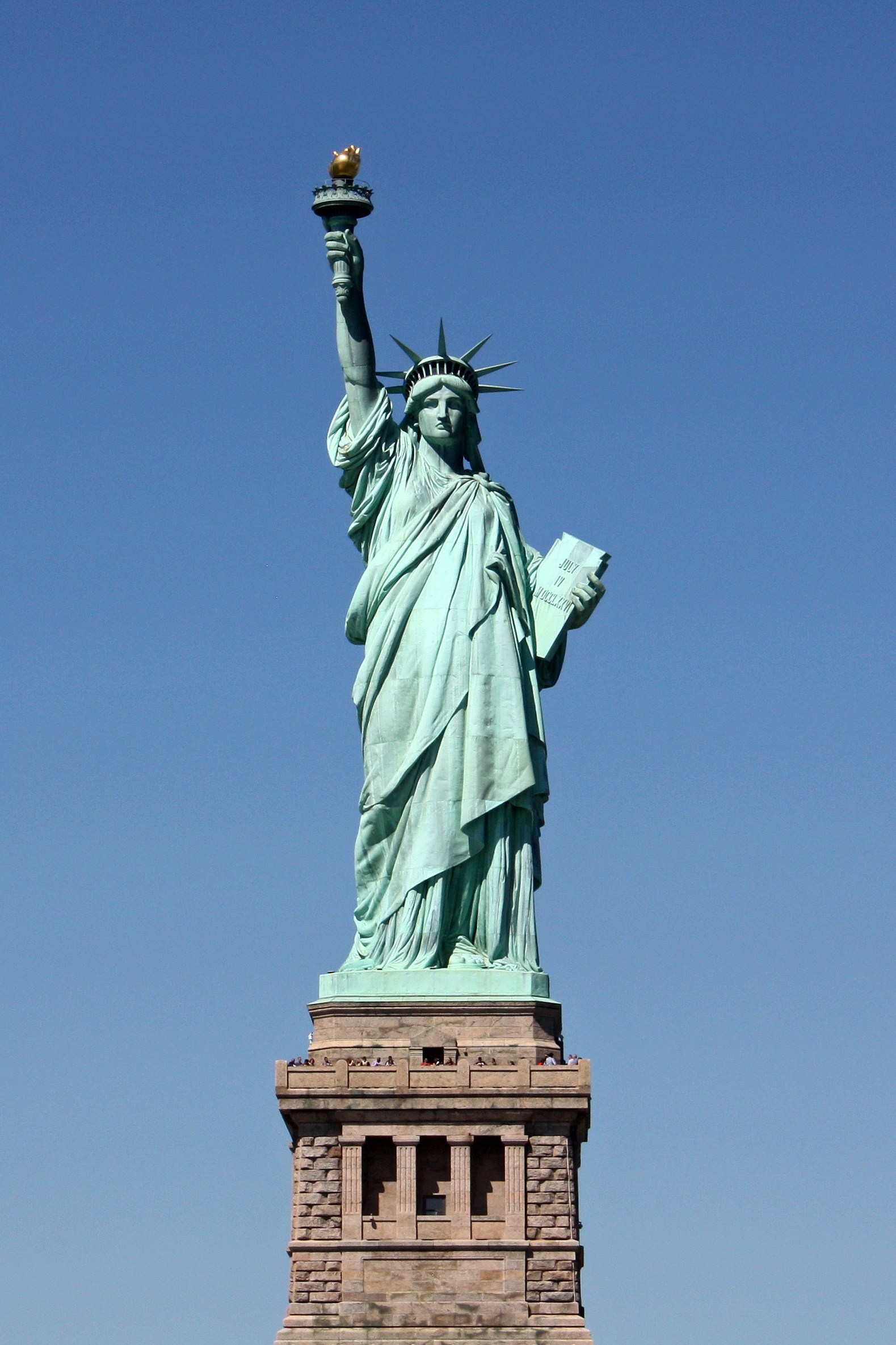 The green Statue of Liberty standing over New York Harbor, holding a torch toward a clear blue sky.