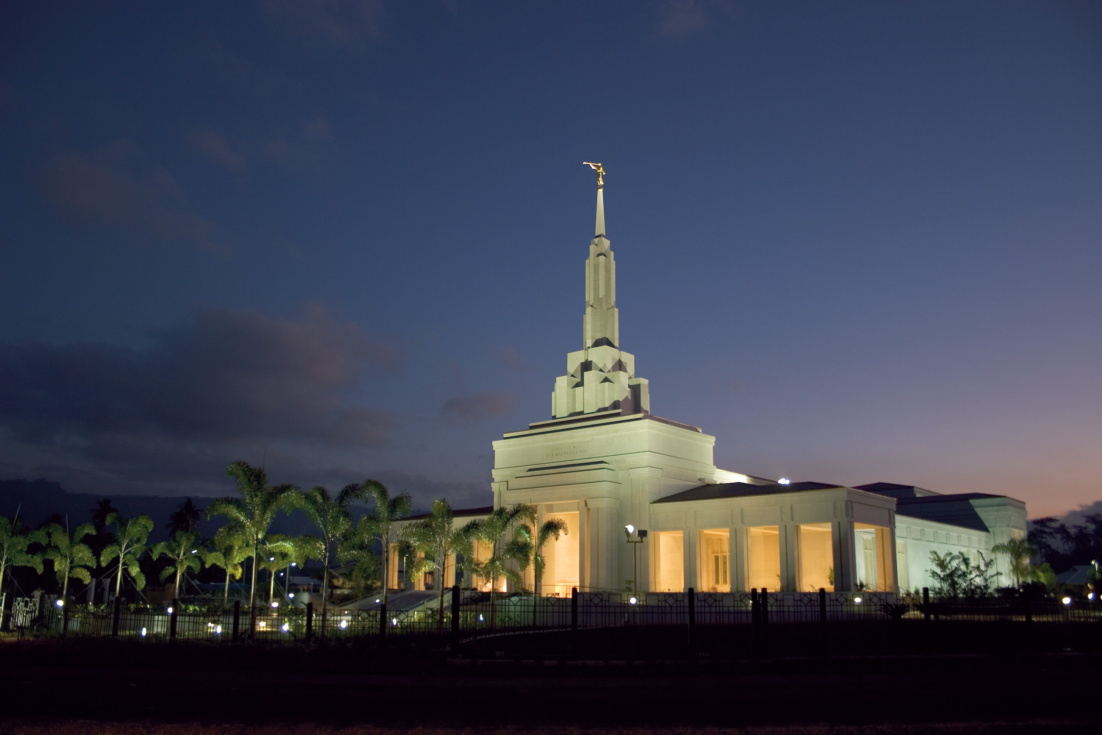 The Apia Samoa Temple lit up at twilight with a row of palm trees leading up to the front door.