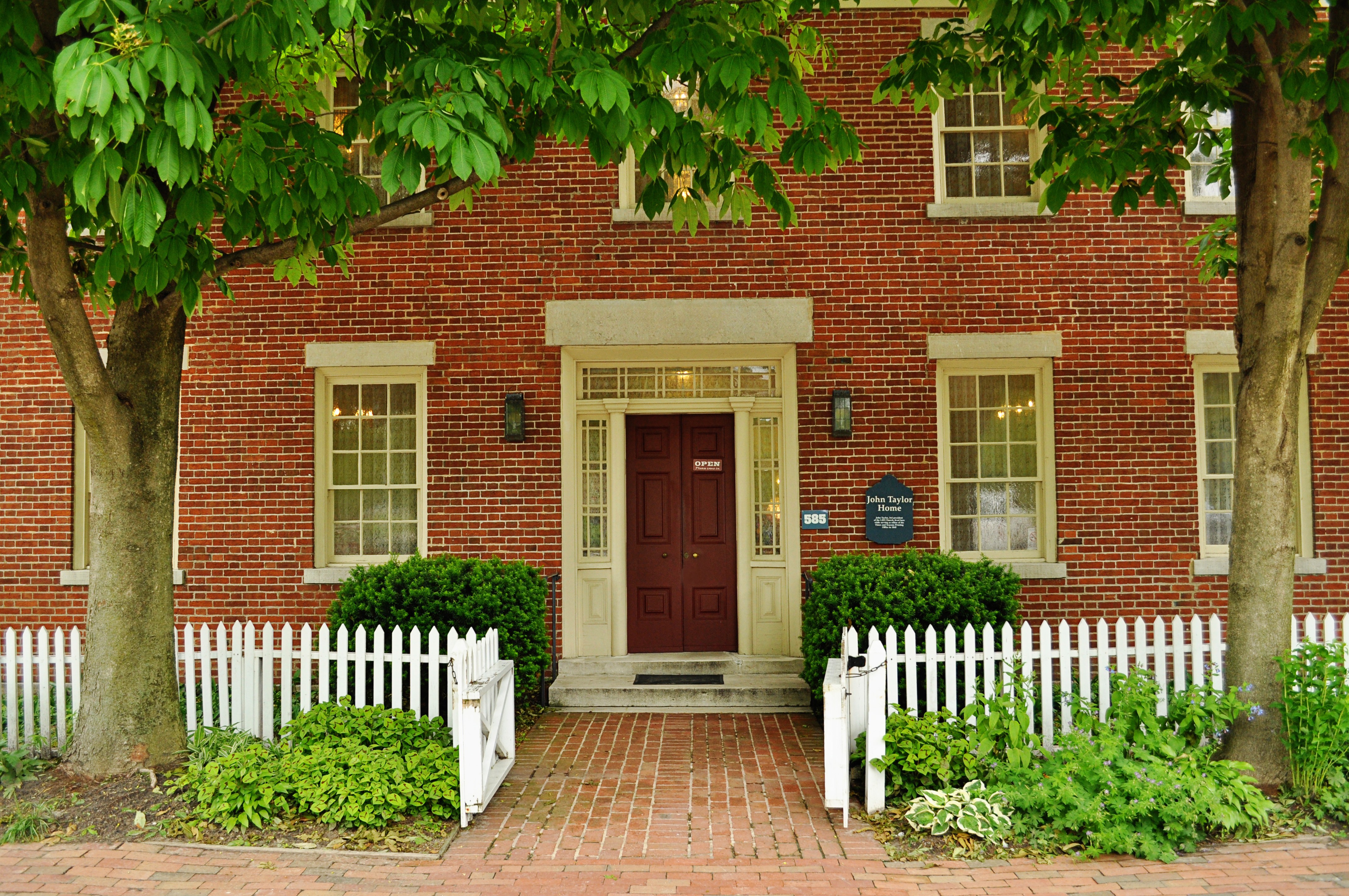 The red-brick home of John Taylor with a white picket fence in front and two trees on either side of the front door.