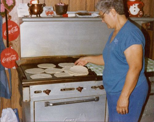 Color photo of an older woman cooking tortillas or some kind of flat bread on a stove top.