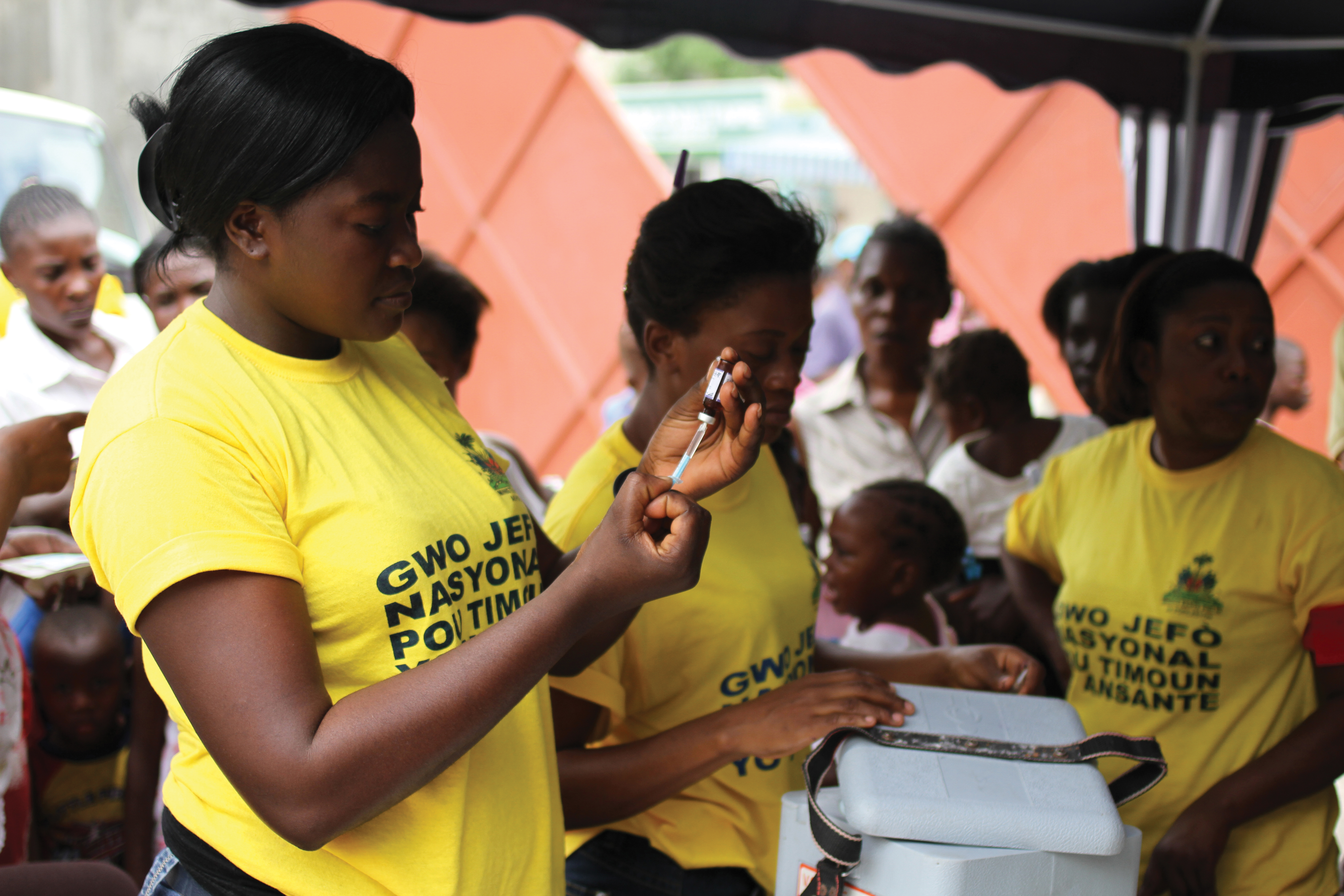 A Haitian woman in a yellow shirt holding a vaccine bottle upside down and filling a syringe while standing near other women.
