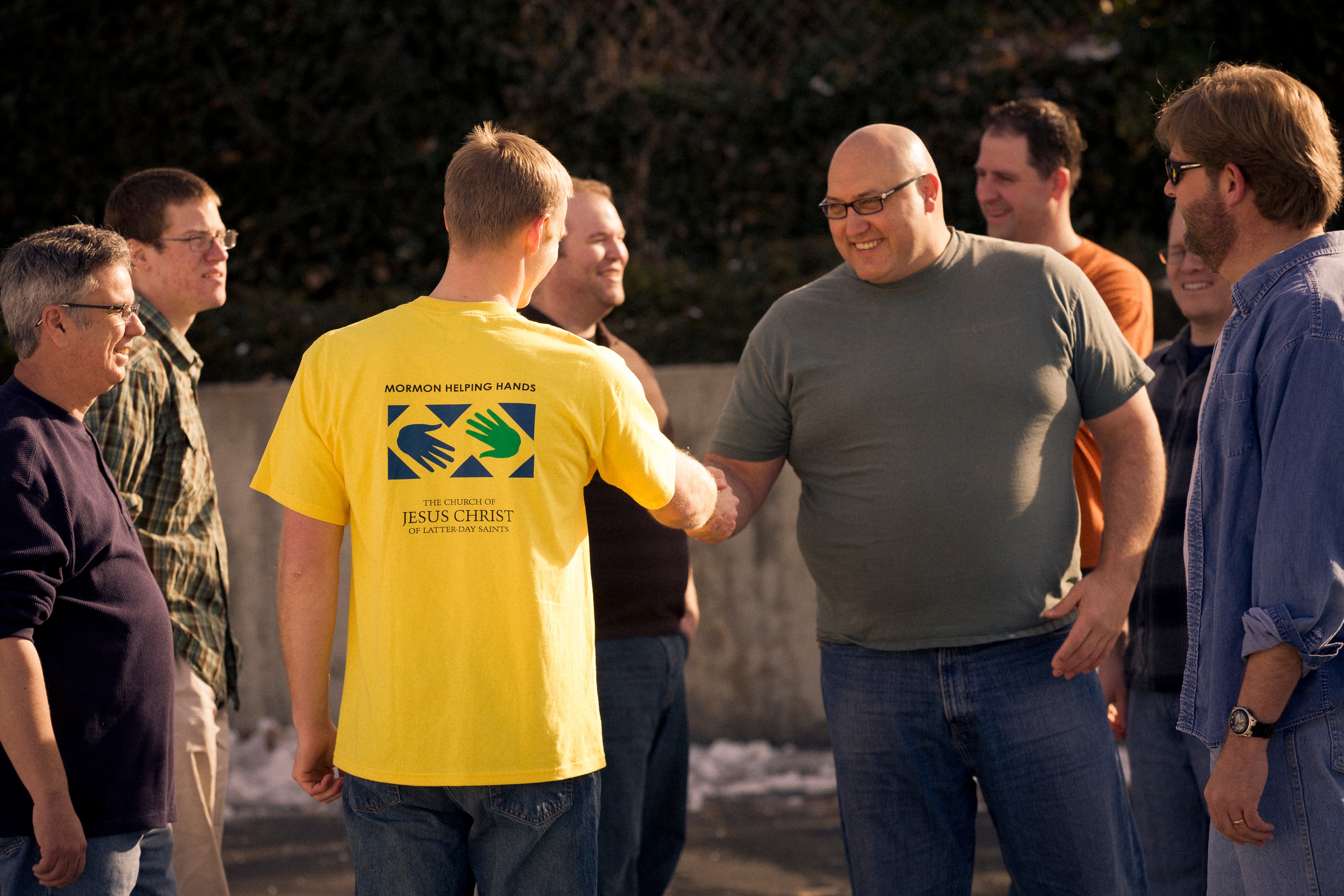 A young adult man in a yellow Mormon Helping Hands tee-shirt is shaking hands with another man.  There are other men standing nearby.