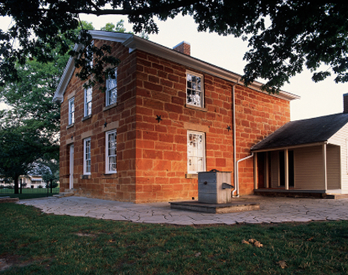 Two-story, sandstone jail building with a well in the foreground, pictured at dusk.
