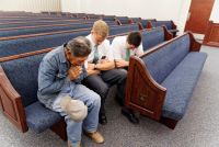 Two elder missionaries sit with a man in a chapel pew and pray