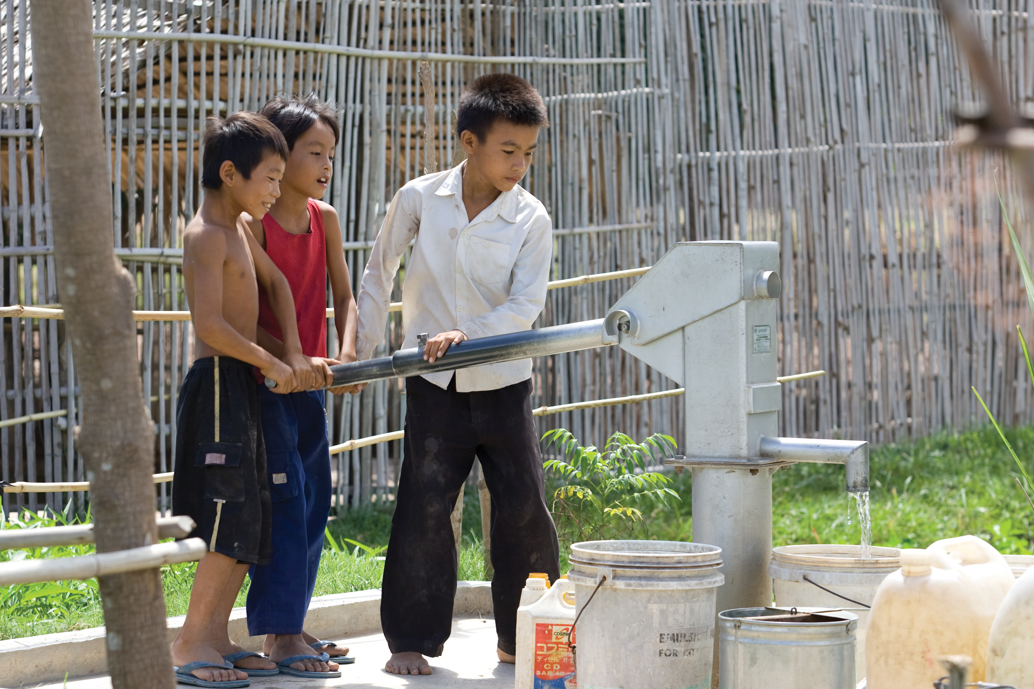 Three young boys in Cambodia standing and pumping clean water into a bucket from a metal hand-operated pump.