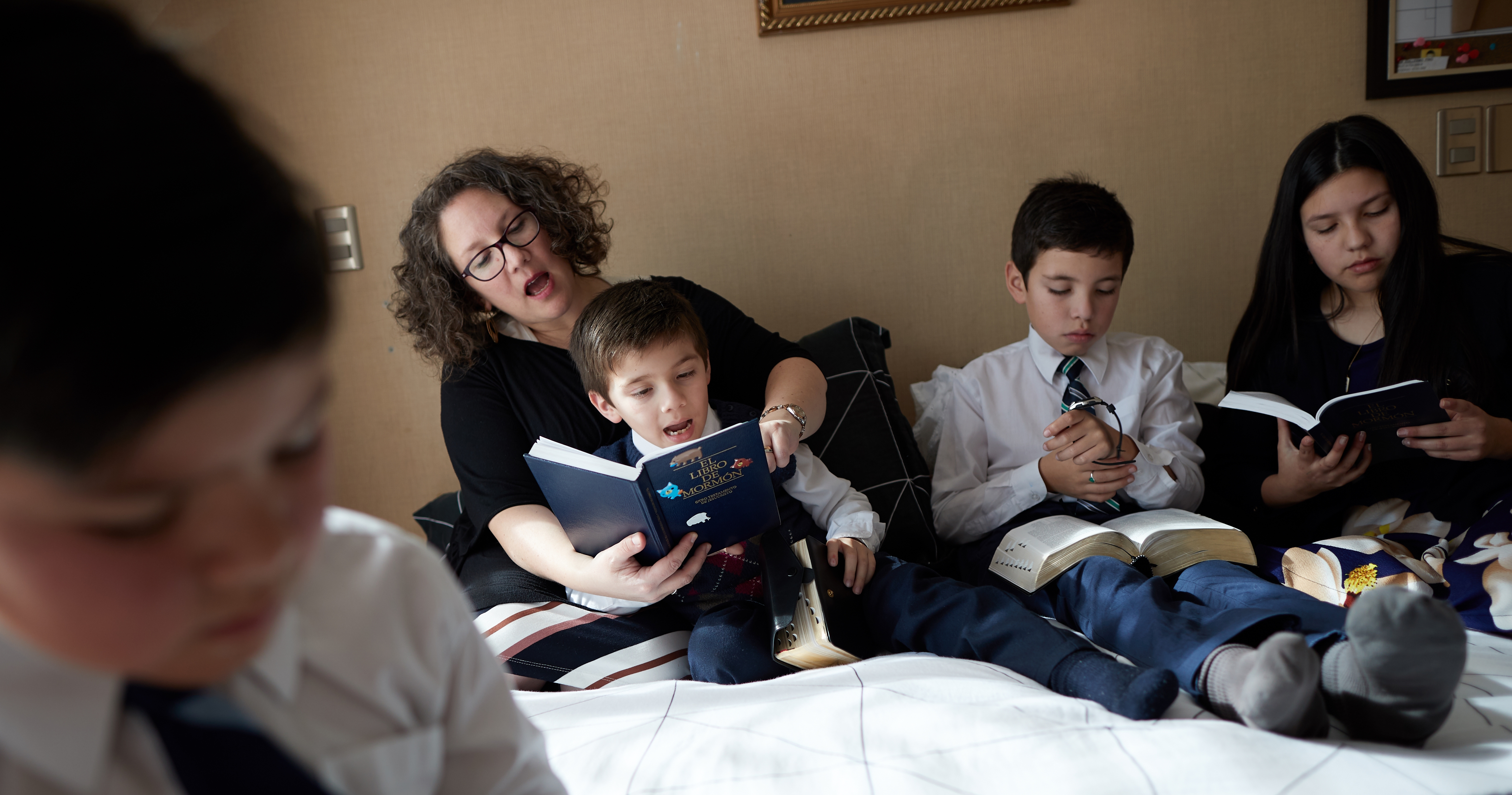 A family sits together on a large bed and reads the scriptures together.