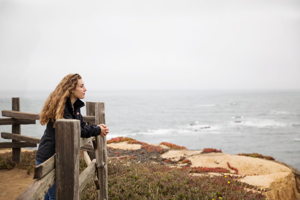 Uma mulher inclina-se em um parapeito à beira-mar contemplando o evangelho de Jesus Cristo