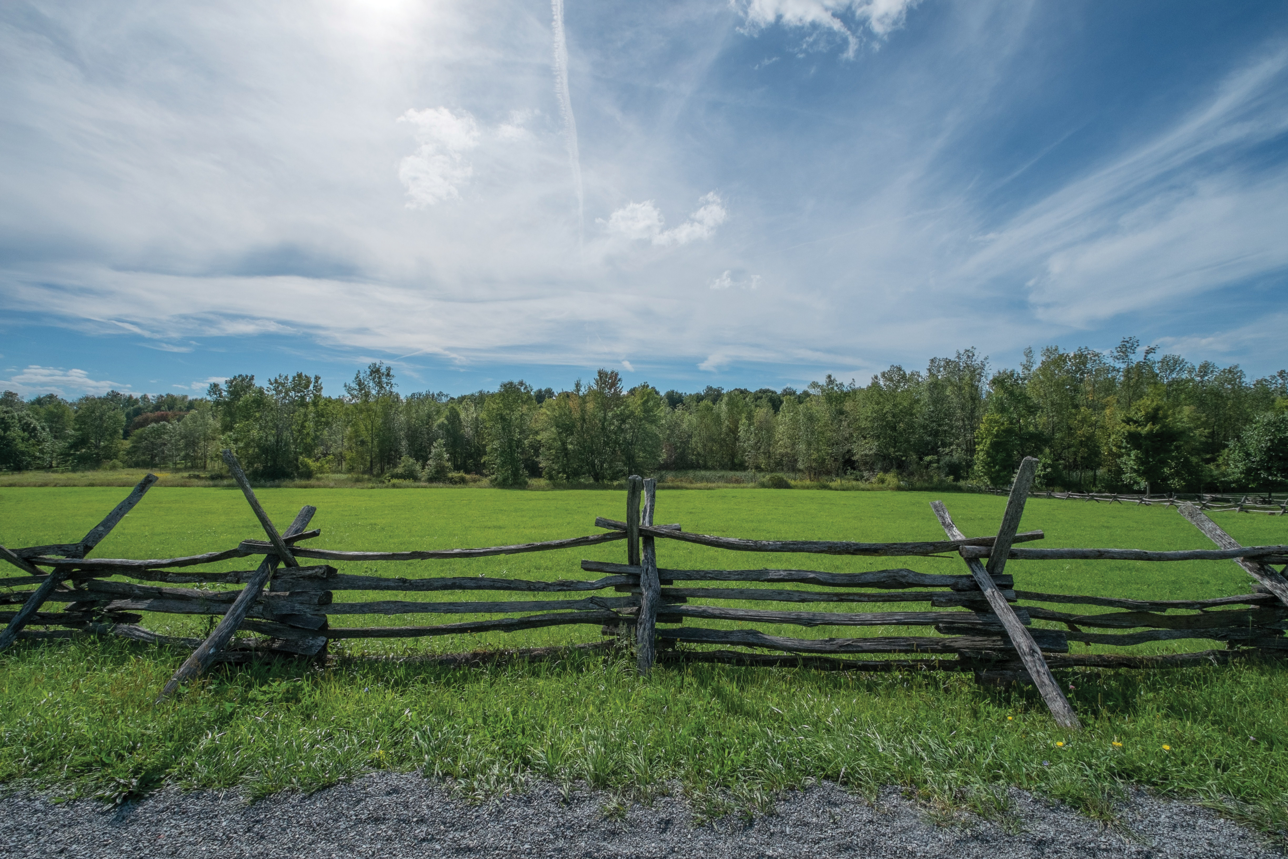 A wooden fence on the Smith family farm, running along the edge of a gravel road, with a green field and trees in the background.
